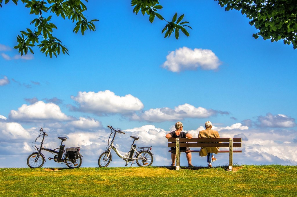 Deux personnes assises sur un banc dans un parc, regardant le ciel bleu parsemé de nuages blanches. À côté, deux vélos sont stationnés sur l'herbe verte.